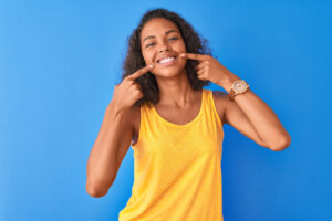 brown woman in a yellow tanktop points to her smile after fixing her teeth with dental bonding