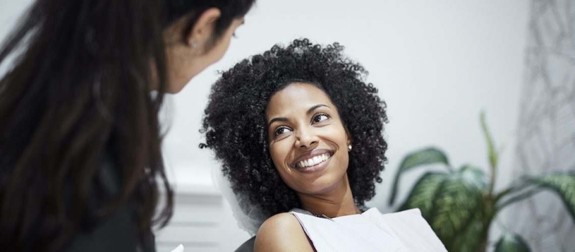 Smiling Black woman with curly hair and a white bib talks to her dentist at her routine appointment