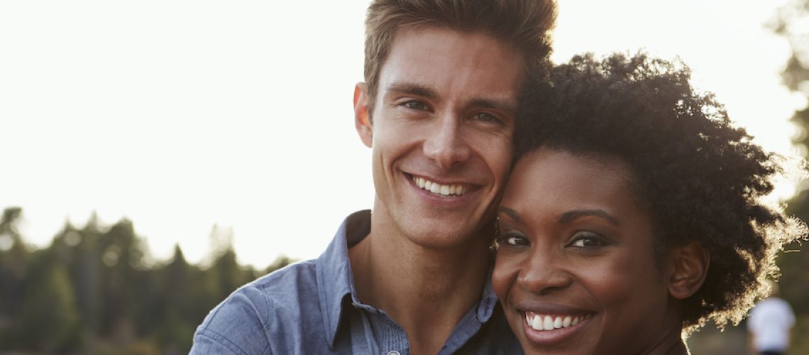 Black and white couple smile as they embrace outside in nature