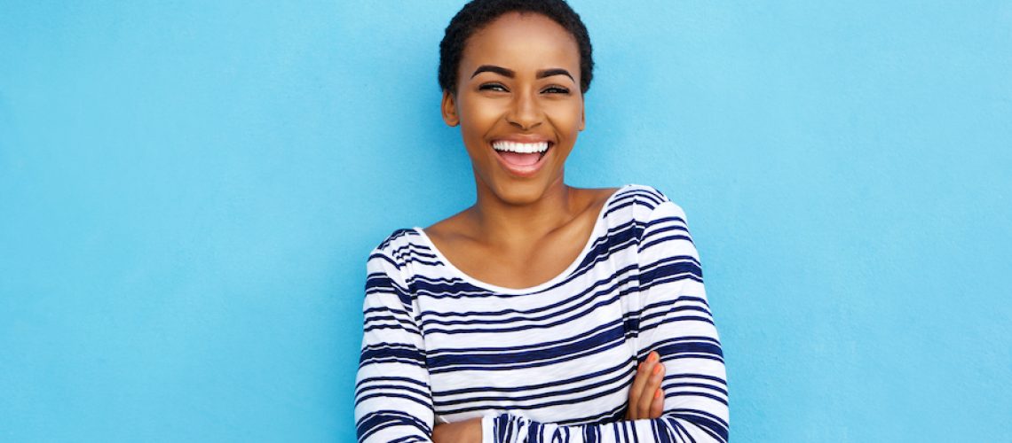 black woman in a striped shirt against a blue wall smiles after cosmetic dentistry
