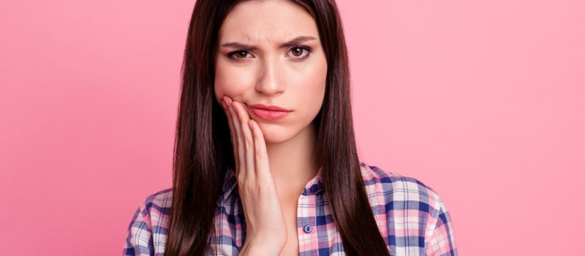 Brunette woman in a plaid shirt cringes in pain and touches her cheek due to bruising after root canal therapy